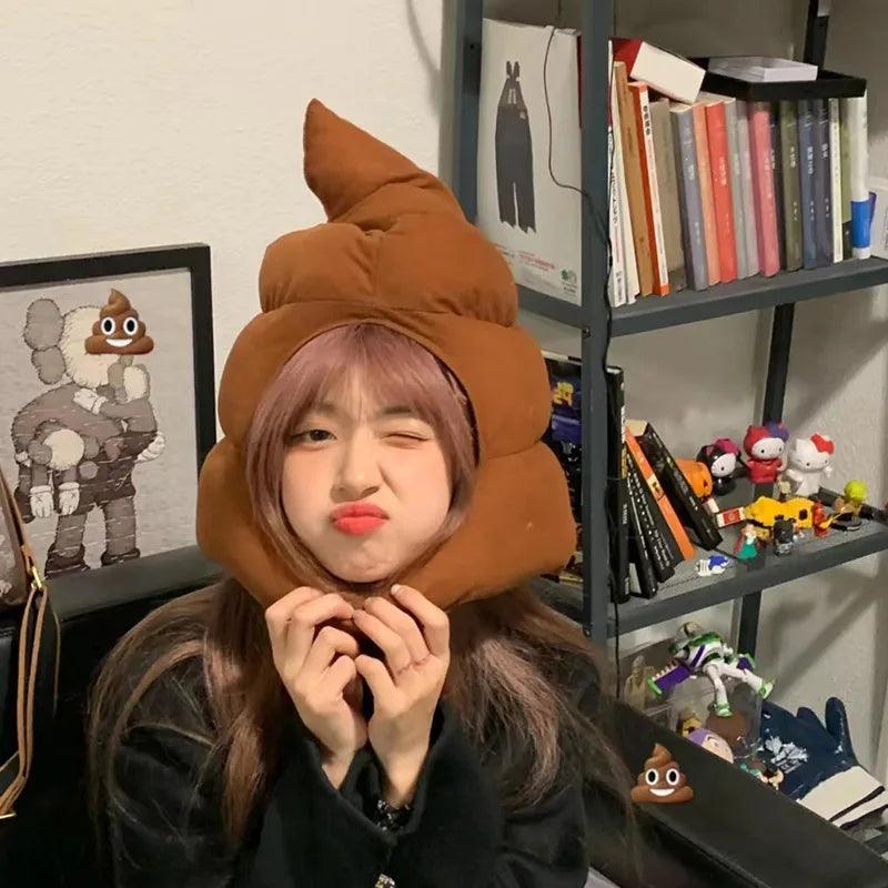 Person wearing a brown poop emoji plush hat indoors with bookshelves, cute poop head hat
