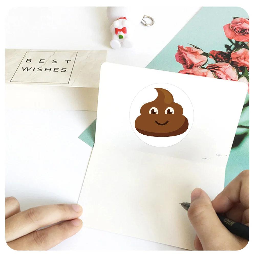 Card with a happy poop emoji sticker, being written on, next to a "Best Wishes" envelope and roses.