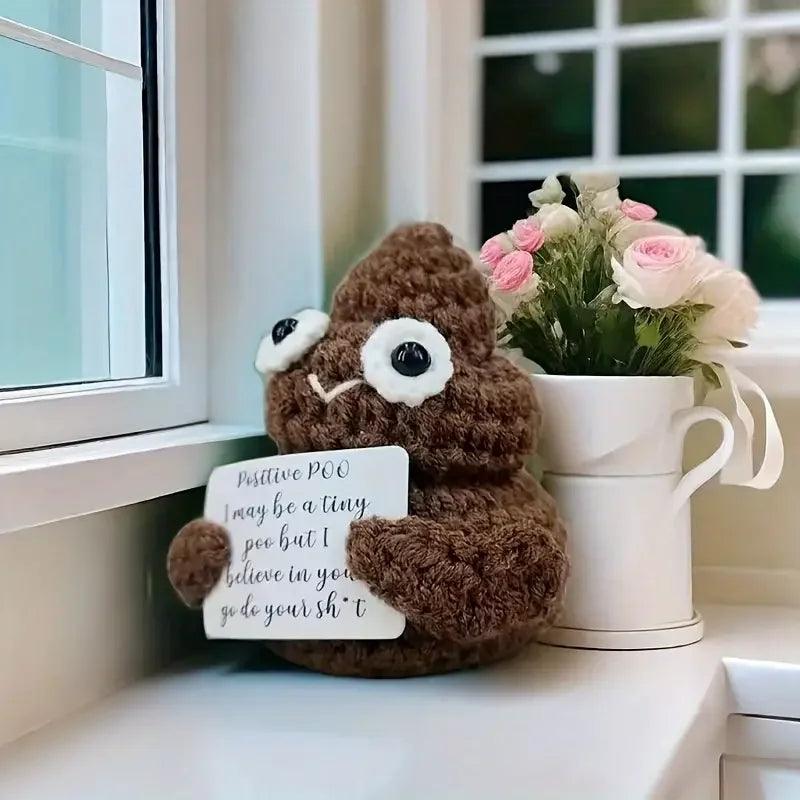 Positive Poop plush next to pink roses in a vase, holding a motivational sign by a window.