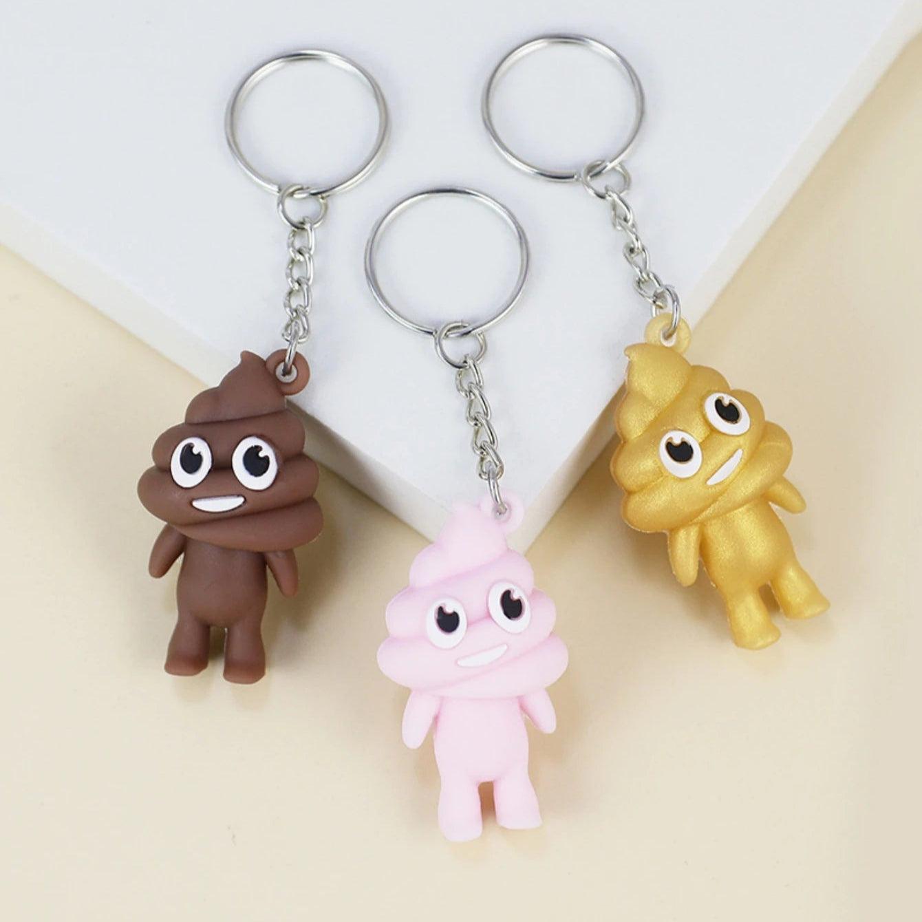 Three poop emoji keychains in brown, pink, and yellow on a surface, each with a metal keyring.